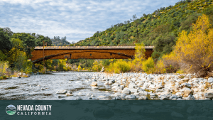 A historic covered wooden bridge spans a rocky river, surrounded by autumn-colored trees and green hills under a partly cloudy sky in Nevada County, California. A banner in the corner proudly reads, "Nevada County.