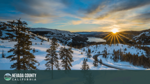 A snowy mountain landscape at sunset, with a winding road and trees in the foreground, and a lake and hills in the distance captures the serene beauty of Nevada County. "Nevada County California" logo appears at the bottom of the image.