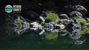A calm river reflects rocks and lush green foliage along its bank, capturing the natural beauty of Nevada County, with the Nevada County, California logo in the upper left corner.