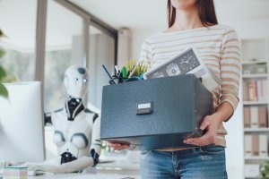 A person in a striped shirt is holding a box filled with office supplies, including a clock, pens, and a potted plant. In the background, a humanoid robot is seated at a desk with a computer in a modern office setting, focused on its work.