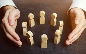 A pair of hands, palms facing down, is positioned around a circle of small wooden figurines standing upright on a dark surface. The light shines from above, casting soft shadows and creating a warm atmosphere reminiscent of quiet labor.
