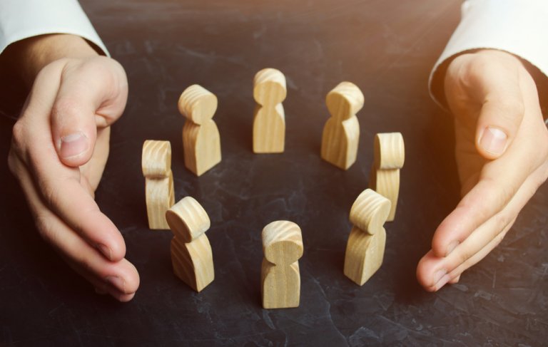 A pair of hands, palms facing down, is positioned around a circle of small wooden figurines standing upright on a dark surface. The light shines from above, casting soft shadows and creating a warm atmosphere reminiscent of quiet labor.