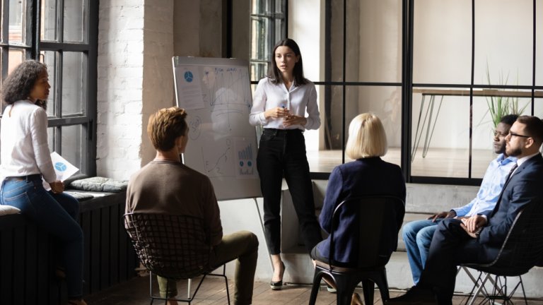 A woman stands next to a flip chart, demonstrating her leadership skills to a small group of seated colleagues in a modern office space. The group of five people listens attentively, with large windows and a brick wall in the background.