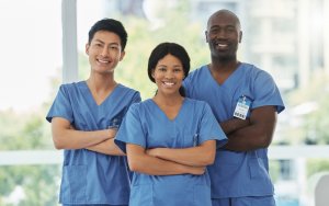Three healthcare professionals in blue scrubs, two men and one woman, stand side by side with arms crossed, smiling at the camera. They are in a brightly lit room with large windows, set against a blurred background that showcases greenery and buildings.