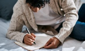 A man in a casual suit writing in a notebook on top of a bed