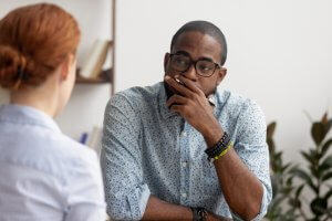 A man wearing glasses and a patterned shirt sits thoughtfully with his hand on his chin, listening to a woman with red hair who is speaking about her job. They are in a well-lit room with bookshelves and plants in the background.