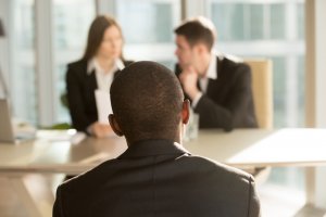 A man sits with his back to the camera, facing two people across a table in what appears to be a job interview setting. The two individuals, dressed in business attire, are engaged in conversation with the man and are holding documents.