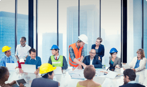 A diverse group of professionals, including construction workers wearing hard hats and office staff, gather around a table in a modern conference room with large windows overlooking a cityscape, collaborating on a project with blueprints and laptops.