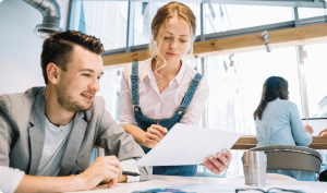 Two people are collaborating in a bright, modern office. A man in a suit jacket sits and looks at a document while a woman in a white shirt and denim overalls stands next to him, holding the paper and pointing at something. Another person works at a desk in the background.