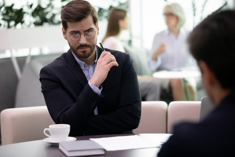 A man in a suit, wearing glasses, is sitting at a table in a professional setting. He is holding a pen and looking focused, as if discussing an important job matter. In the background, two people are engaged in conversation. A cup of coffee and a notebook are on the table.