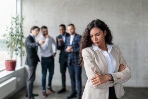 A woman stands with her arms crossed, looking down and to the side, with a serious expression. In the background, four men in business attire are engaged in conversation and looking at a phone, discussing work. The setting appears to be a modern office space.
