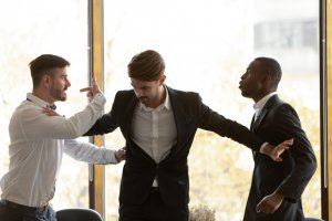 Three men in a work setting having a heated argument. The man in the middle appears to be trying to break up the confrontation, placing a hand on each individual's chest to separate them. The environment has large windows with blurred outdoor scenery.