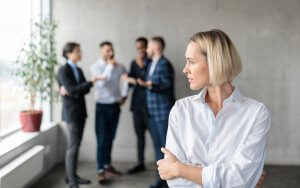A woman with short blonde hair and a white shirt stands in the foreground with her arms crossed, looking to the side. In the blurred background, four coworkers are engaged in a conversation near a large window and a potted plant.