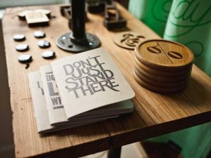 A wooden table displays small notebooks, one with "DON'T JUST STAND THERE" on the cover, wooden coasters, and several round pins. Bright green fabric hangs in the background at a City of Albany event.