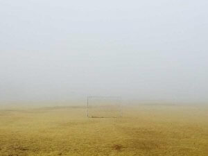 A single soccer goal stands on a yellowish, empty field in the City of Albany, surrounded by thick fog that obscures the background and creates a sense of emptiness and isolation.