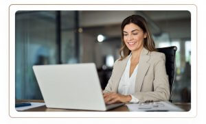 A woman in formal attire sitting while smiling and navigating her laptop