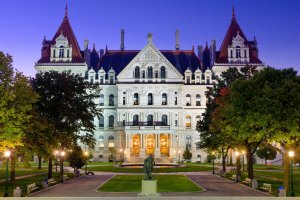 The New York State Capitol building in the City of Albany is illuminated at dusk, with a statue in the foreground, green lawns, and trees framing the scene.