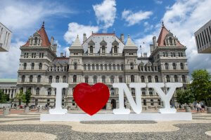 A large “I ♥ NY” sign stands in front of the historic New York State Capitol building in the City of Albany, featuring ornate architecture and pointed roofs under a partly cloudy sky.