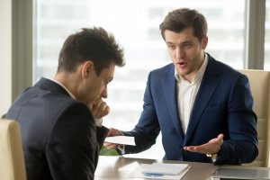 Two men in business attire are having a serious discussion in an office. One man, with a concerned expression, holds a document, while the other man, appearing frustrated, gestures with his hands. There is a notebook and pen on the table between them as they debate their sales leads.