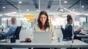 A woman is smiling and working on a laptop in a modern, open-plan office. She is seated at a desk with a lamp, a coffee cup, and a few documents. In the background, two colleagues are also working at their desks, with colorful sticky notes on the glass windows—a vibrant scene of productive employment.