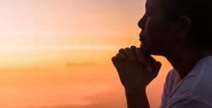 Silhouetted person with hands clasped and head resting on hands, sitting against a warm, colorful sunset sky. The scene evokes a sense of peace, contemplation, and tranquility. A light-colored shirt hints at their role in public service as they look towards the horizon.