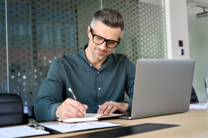 A man with glasses and gray hair, wearing a green button-up shirt, is sitting at a desk in a modern office. He is writing in a notebook with his right hand while using a laptop for his remote work. The background features a glass wall with a dot pattern.