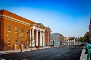 A street view of a quiet, small-town Main Street with a prominent brick building featuring large white columns. The street is lined with lampposts, American flags, and various storefronts, showcasing the town's charm and talent for creating a picturesque, inviting atmosphere under a clear blue sky.