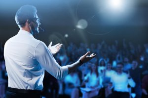A man wearing a white shirt and glasses stands on stage with a headset microphone, exuding leadership as he gestures with open hands while facing an attentive audience. The background is dim with a bright spotlight shining down, highlighting the speaker's presence.