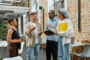 A group of four people are standing and talking in a modern indoor setting with exposed brick walls. They are holding notebooks and documents, engaging in a lively discussion about recent governmental policies. The space around them has tables and chairs, indicating a casual meeting environment.