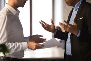 Two people in a discussion, one holding a document. The person on the left is dressed in a white shirt, and the person on the right, who leads the conversation, is wearing a black blazer over a blue shirt. Both are gesturing with their hands. The background is blurred with daylight visible through windows.