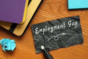 A black, crumpled piece of paper with the words "Employment Gap" written in white text and an arrow below it lies on a wooden surface. Nearby are notebooks and a crumpled blue paper ball, along with a marker capped with a white tip—symbolizing employment challenges.