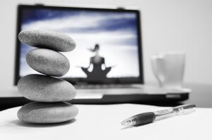 A stack of four balanced stones sits on a white surface in front of a laptop showing an image of a person meditating under a blue sky. Representing the calm and balance often sought by those in public service, a pen and white mug are also visible in the background.