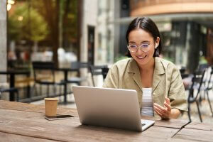 A person with short hair and glasses is smiling while remotely working on a laptop at an outdoor cafe. They are wearing a beige shirt, and there is a smartphone and a disposable coffee cup on the wooden table. The background shows blurry cafe chairs and greenery.