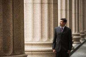 A man in a dark suit and striped tie stands beside tall, stone columns, exuding an air of talent. He is looking to his right with a slight smile. The columns are part of an outdoor architectural setting, suggesting a formal or professional environment.