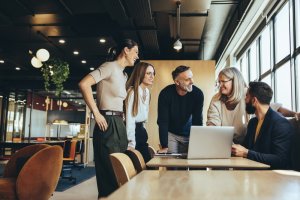 A group of five people in casual business attire are gathered around a laptop in a modern office. They are smiling and engaged in conversation, embodying the spirit of teamwork and collaboration. The office has large windows, letting in natural light, and is stylishly furnished.