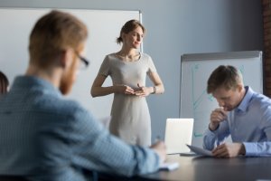 A woman leads a discussion, addressing three colleagues in a meeting room. She stands next to a whiteboard and a flip chart while the others sit at a table with notebooks and papers in front of them. One person writes, while another appears to be deep in thought.