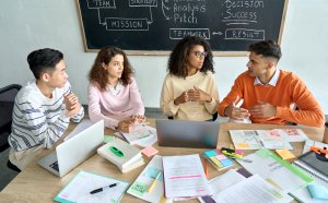 A diverse group of four people are having a discussion at a meeting table cluttered with laptops, documents, and office supplies. A chalkboard in the background has business-related notes and government diagrams. The atmosphere appears collaborative and focused.