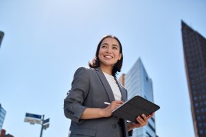 A woman dressed in a gray blazer and white shirt, holding a tablet, smiles while standing outdoors in a cityscape with tall buildings and a clear blue sky in the background, embodying professionalism and modern employment opportunities.
