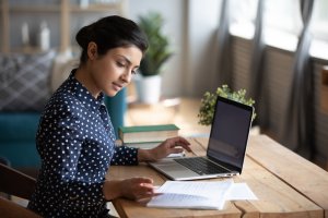 A woman sits at a wooden table, focused on her remote work. She is using a laptop and glancing at a notebook with handwritten notes. Dressed in a navy blue polka dot blouse, she bathes in the light streaming from nearby windows. A small potted plant adds a touch of nature to the scene.