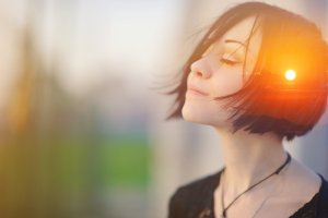 A young woman with short, dark hair stands with her eyes closed and head tilted slightly upward, enjoying the sunlight. She appears calm and content, embodying the tranquility often associated with a dedicated public service worker, basking in the warm glow from the setting sun in the background.