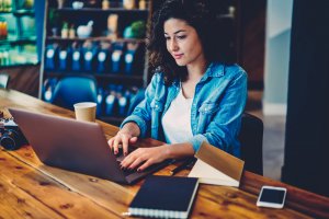A woman with curly hair, wearing a blue denim shirt, works on a laptop at a wooden table. A notebook, a book, a smartphone, and a disposable coffee cup are on the table. The background includes shelves with various items in a cozy indoor setting, giving off an air of quiet concentration as she tackles her government work.