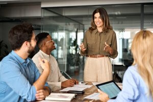 A woman stands at the head of a conference table, exemplifying leadership as she speaks to three seated colleagues. All are engaged and smiling. The woman holds a tablet, and papers and notebooks are spread out on the table. The room features a modern office design with glass walls.