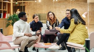 A team of five people engaged in a serious discussion while seated in a modern office space. They are looking at documents and a laptop, with expressions of focus and concentration. The background features glass walls, shelves with books, and minimalistic decor.