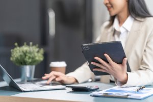 A person in business attire sits at a desk, holding a tablet and using a laptop. There is a potted plant, a takeaway coffee cup, a calculator, and papers on the desk. The background features the sleek design of a modern office building often seen in government institutions.