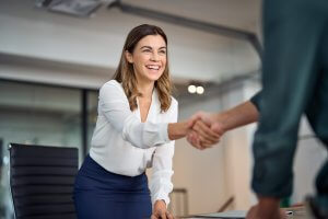 A smiling woman in professional attire leans forward across a table to shake hands with a person whose face is not visible, exuding an air of leadership. The setting appears to be a modern office, with a large window and an empty chair in the background.