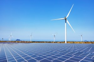 An expansive field of solar panels under a clear blue sky, with several large wind turbines standing tall in the background. The scene is set against distant hills and the ocean, showcasing government investments in renewable energy sources amidst a serene landscape.