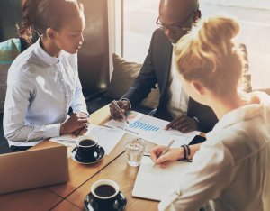 Three people are sitting around a table having a business meeting in the public sector. They are examining charts and graphs on paper. Two cups of coffee, a laptop, and a glass of water are on the table. Sunlight streams in, illuminating the group as they collaborate.