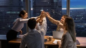 A group of five colleagues, dressed in business attire, are gathered around a table in an office with large windows overlooking a city at dusk. They are high-fiving each other, smiling, and celebrating, suggesting a successful meeting or AI-powered collaboration.