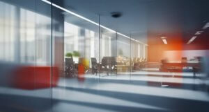 A modern work space with glass walls and partitions, featuring a conference table, chairs, and potted plants. The sunlight streams in through large windows, creating a bright and airy atmosphere. The interior design is sleek, with a mix of red and neutral tones.