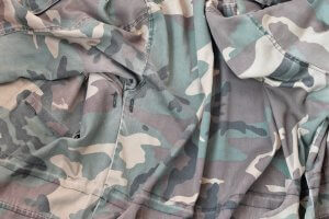 A close-up image of a crumpled, green and brown camouflage-patterned fabric. The fabric appears to be part of a military or outdoor garment, such as a jacket or pants, commonly worn by veterans, featuring various shades of green, brown, and beige in a camouflage design.